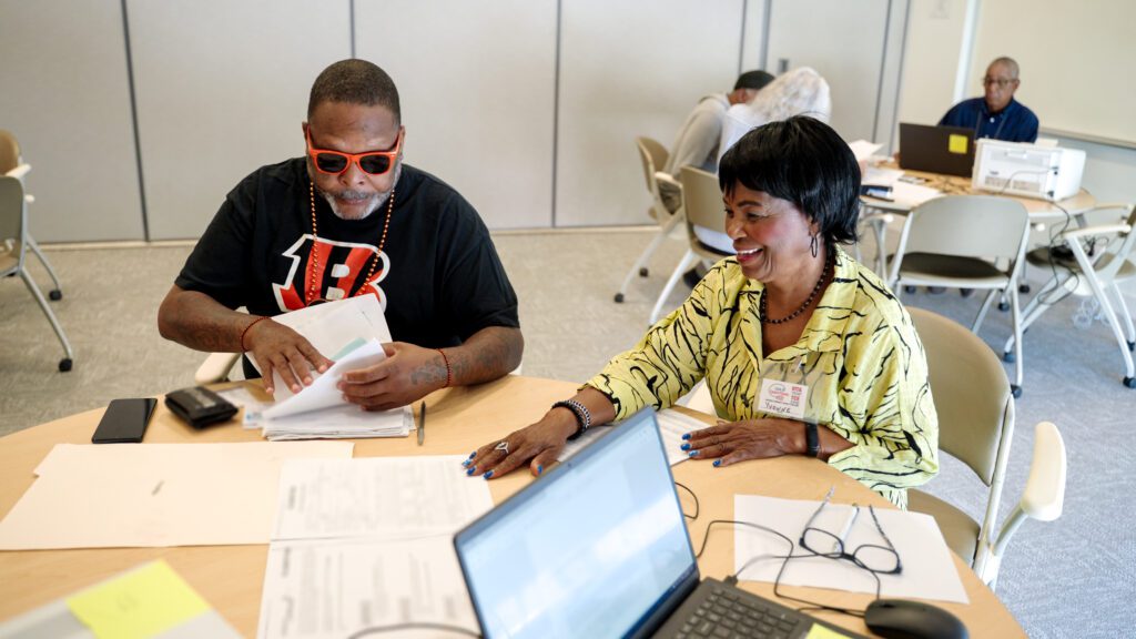 Two people seated at a table reviewing paperwork together at a free tax preparation site, with laptops and other volunteers in the background.